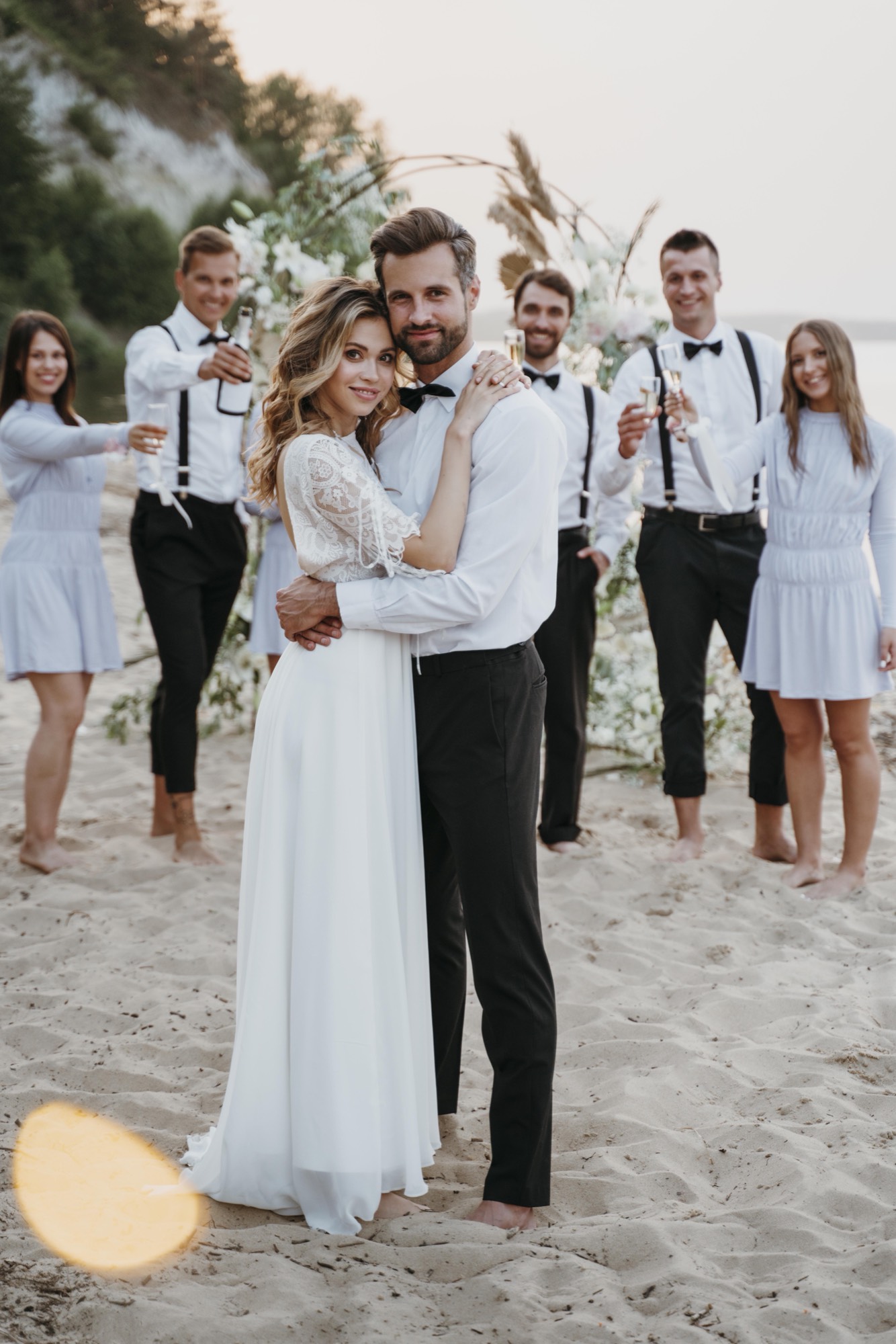 Bride and groom at a beach wedding in Cabo with the wedding party toasting under a floral arch